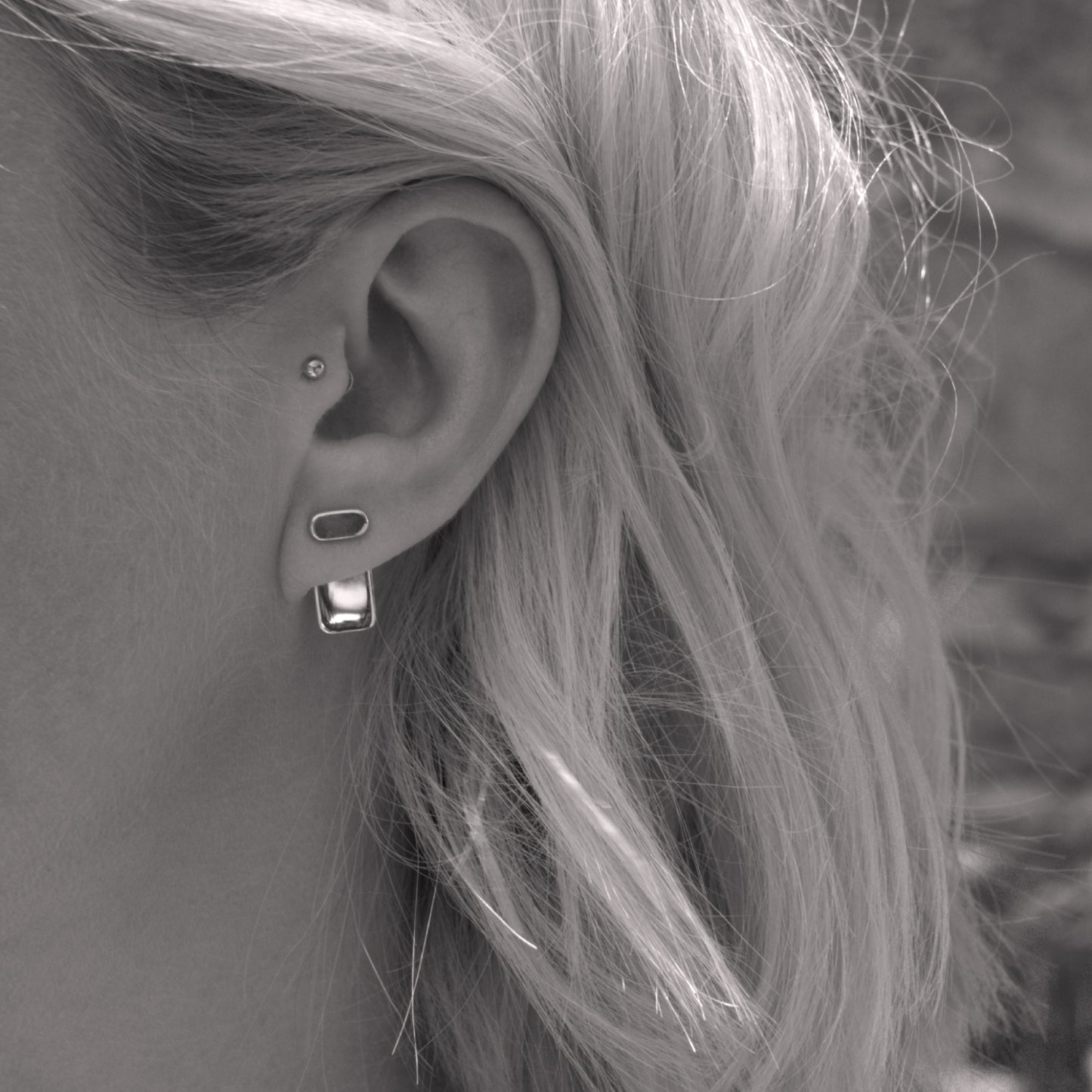 A close-up of a minimalist silver earring in a woman's ear. Black and white.