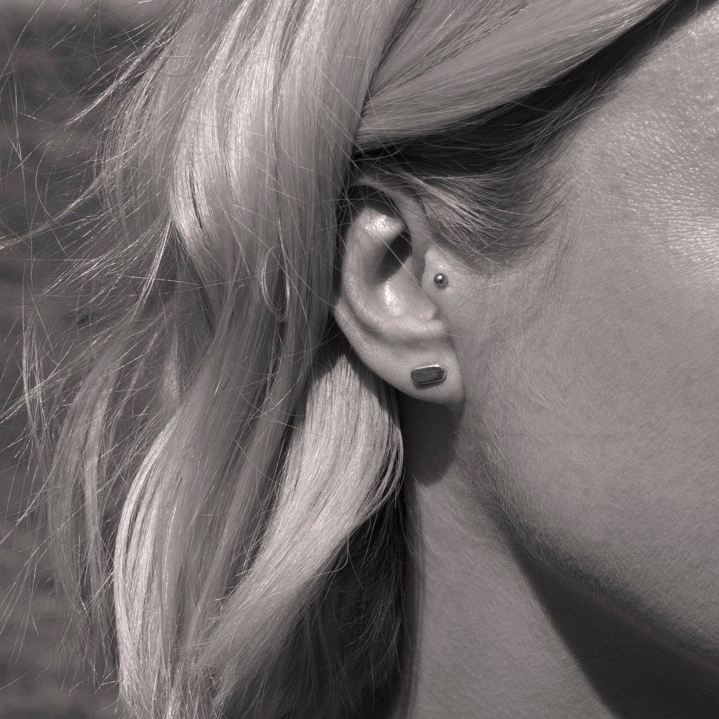 A close-up of a silver earring with a minimalist design in a flat capsule shape in a woman's ear. Black and white.