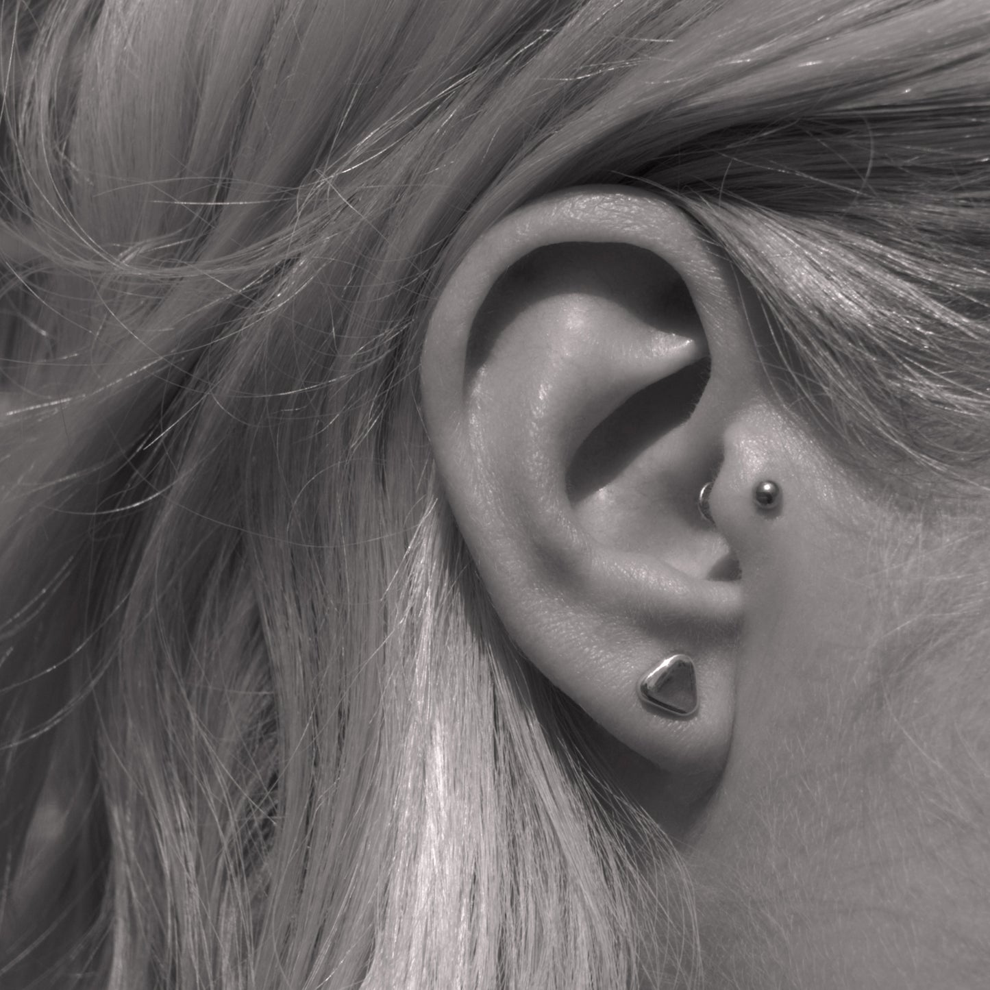 A close-up of a small minimalist design silver earring in a soft triangular shape in a woman's ear. Black and white.