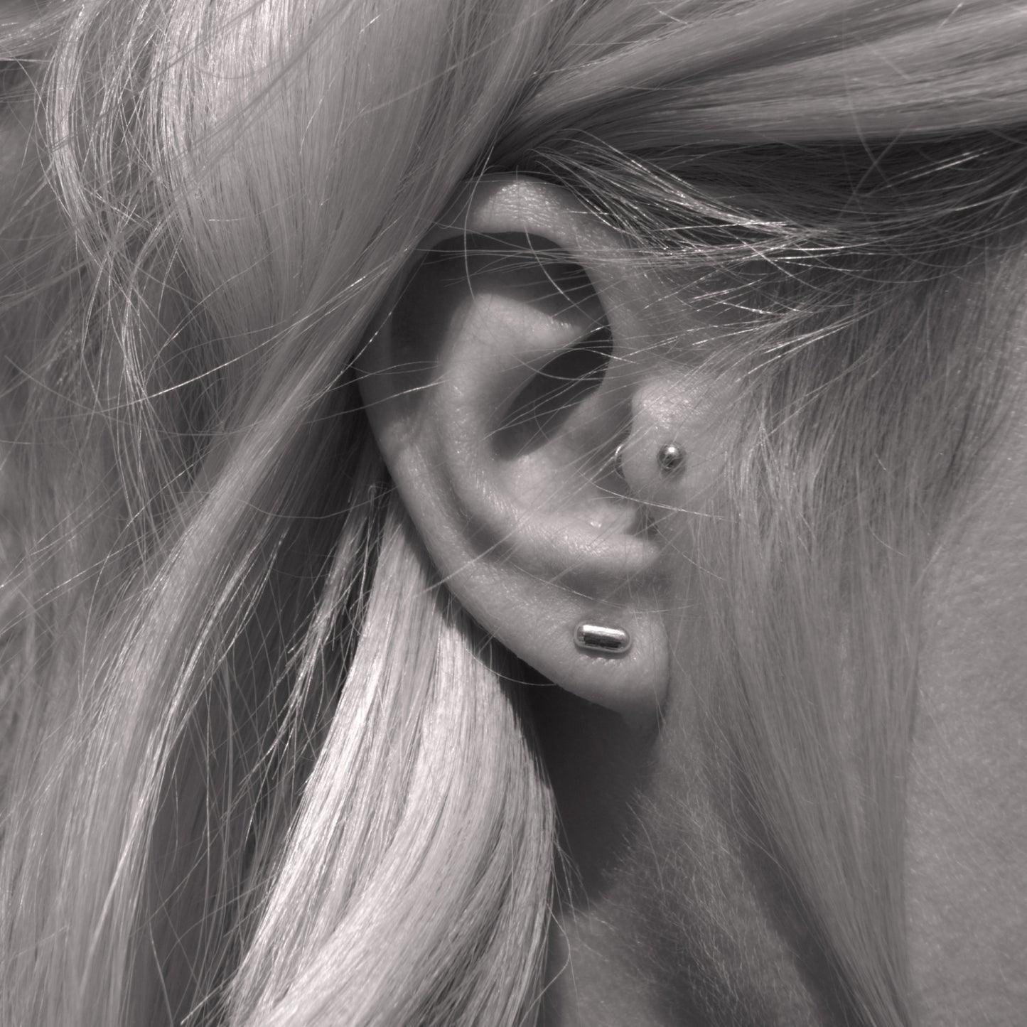 A close-up of a silver earring with a minimalist design in a capsule shape in woman's ear. Black and white.