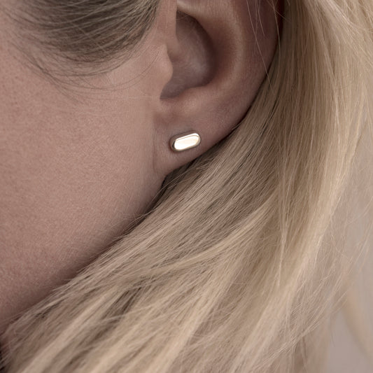 A close-up of a silver earring with a minimalist design in a flat capsule shape in a woman's ear.