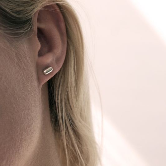 A close-up of a silver earring with a minimalist design in a capsule shape in a woman's ear.