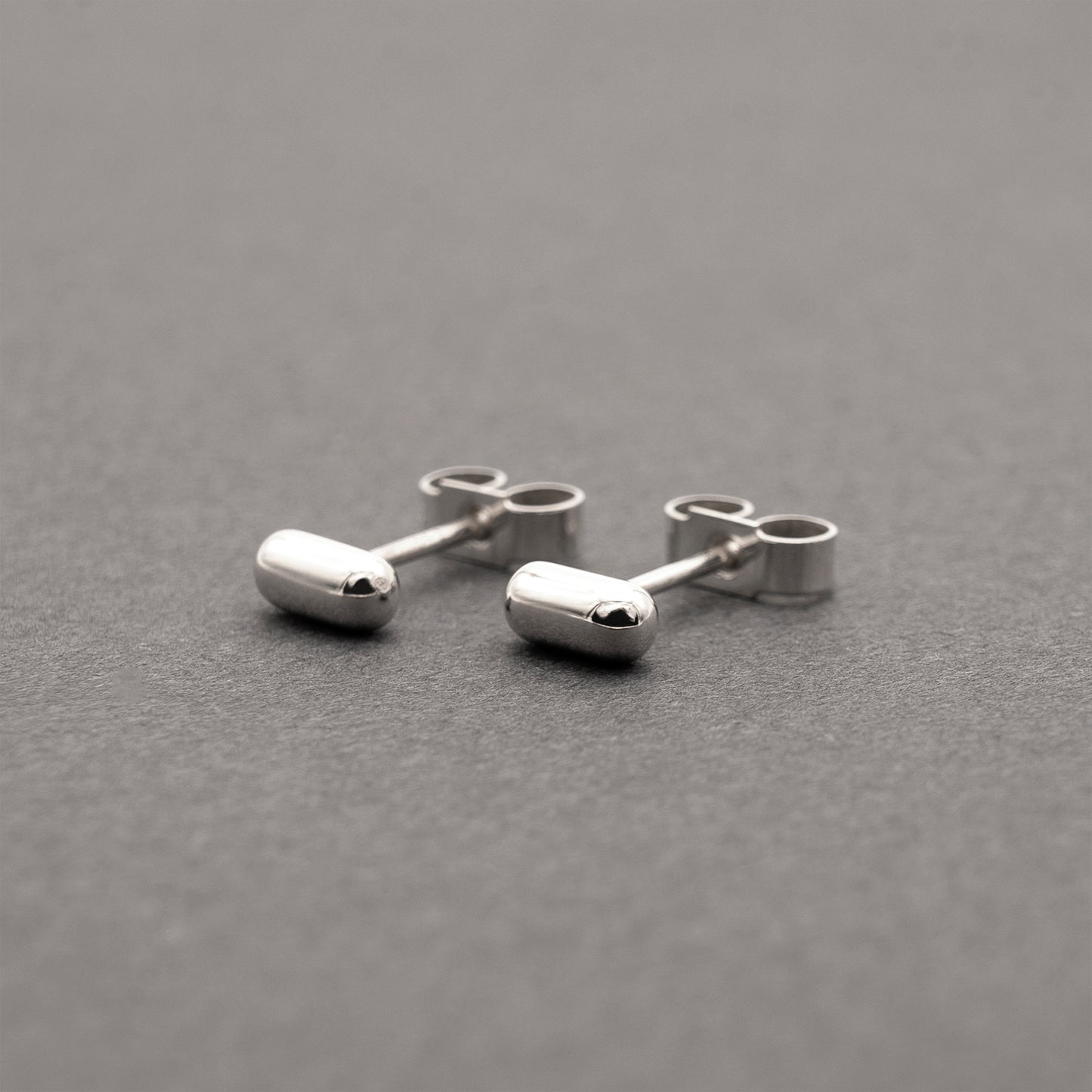A slight side view of two silver earrings with a minimalist design in a capsule shape.
