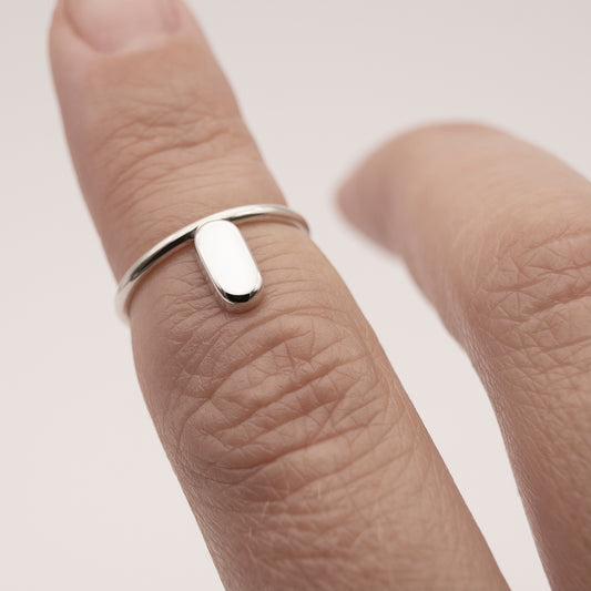 A close-up of a silver ring with a minimalist design and a flat capsule element on a woman's index finger above the knuckle.