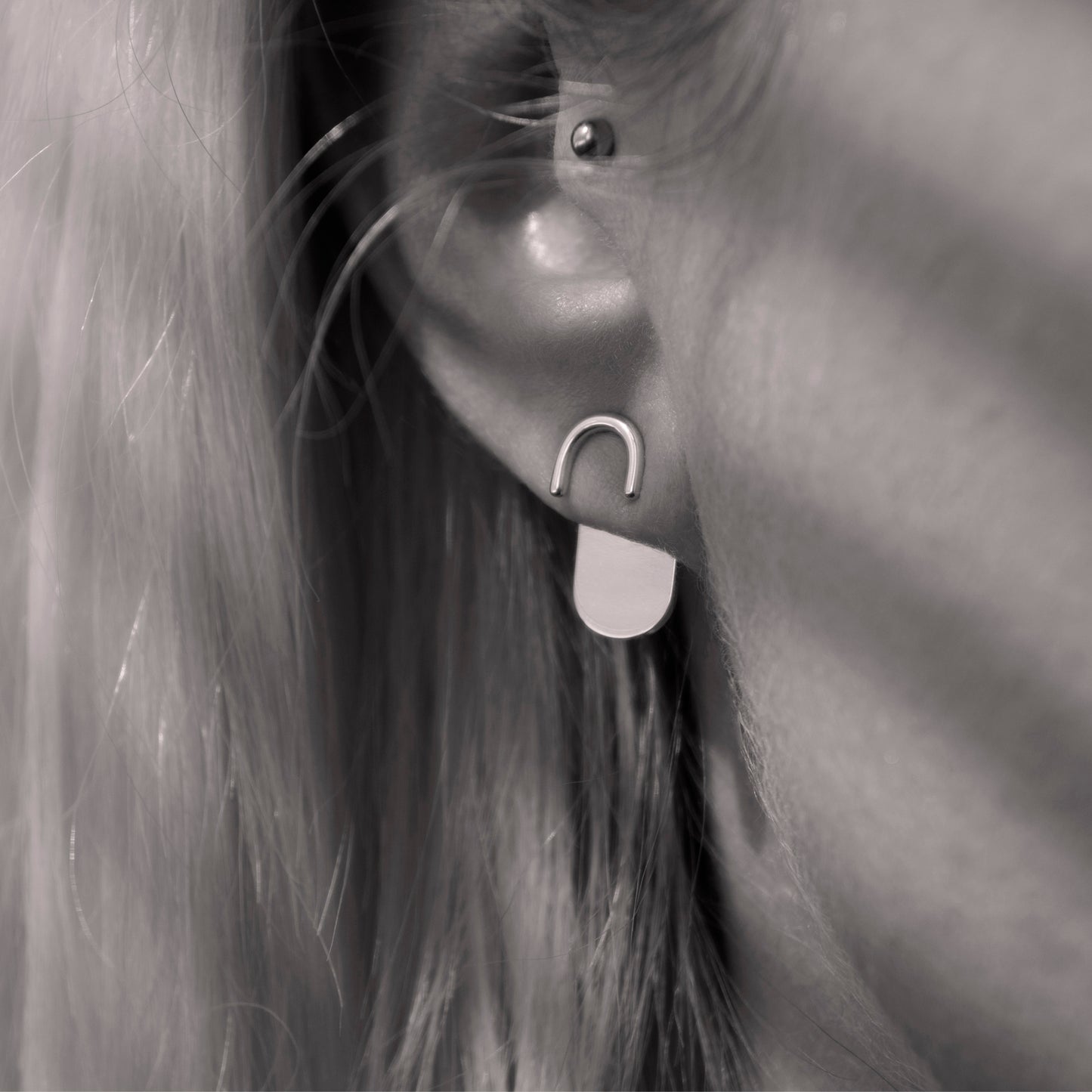 A close-up of a minimalist silver earring in a woman's ear. Black and white.