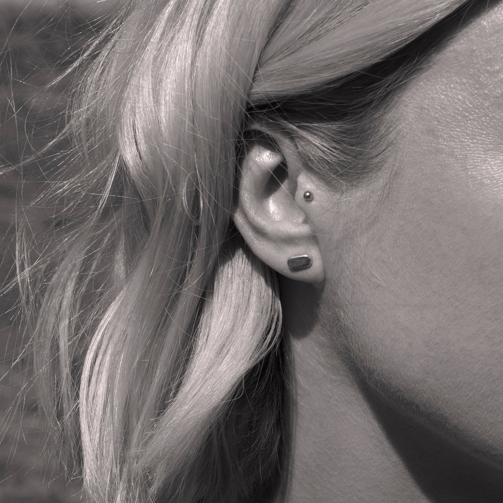 A close-up of a silver earring with a minimalist design in a flat capsule shape in a woman's ear. Black and white.