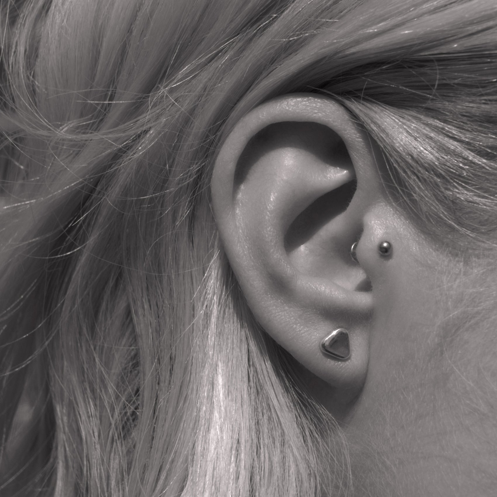 A close-up of a small minimalist design silver earring in a soft triangular shape in a woman's ear. Black and white.