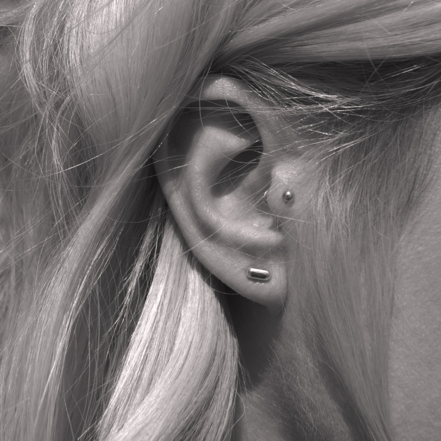 A close-up of a silver earring with a minimalist design in a capsule shape in woman's ear. Black and white.