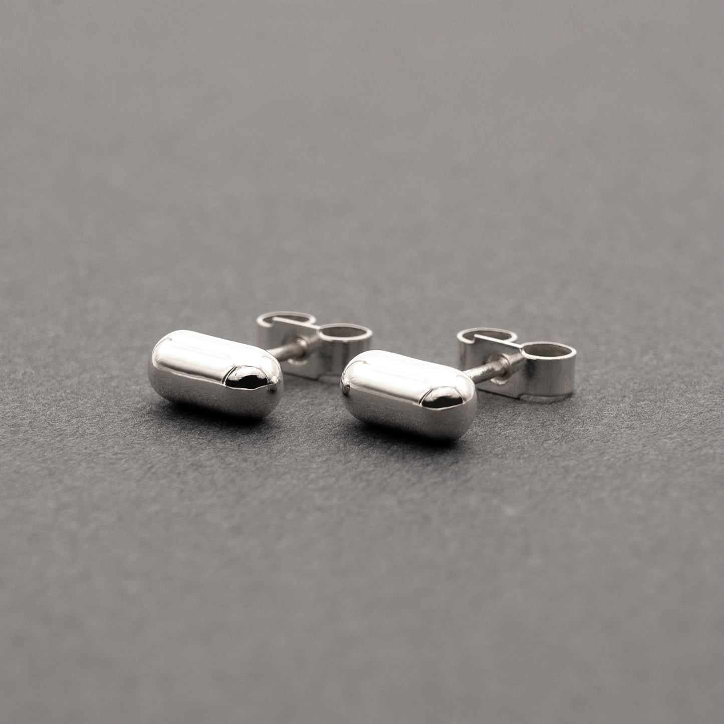 A slight side view of two silver earrings with a minimalist design in a capsule shape.
