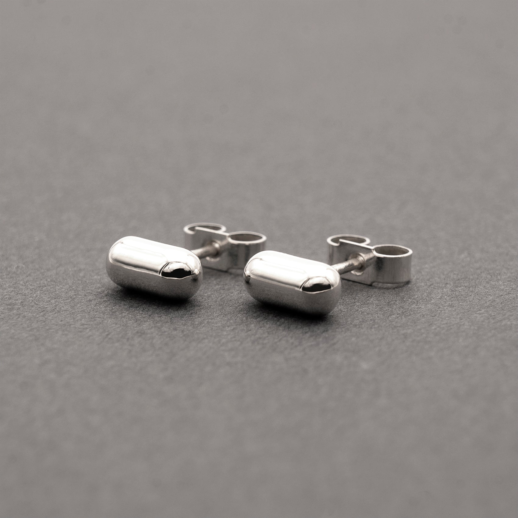 A slight side view of two silver earrings with a minimalist design in a capsule shape.