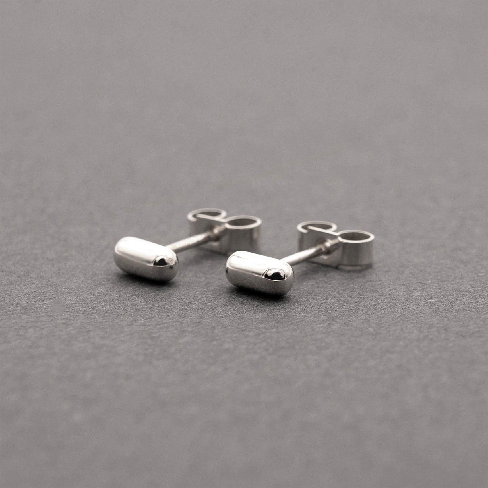 A slight side view of two silver earrings with a minimalist design in a capsule shape.