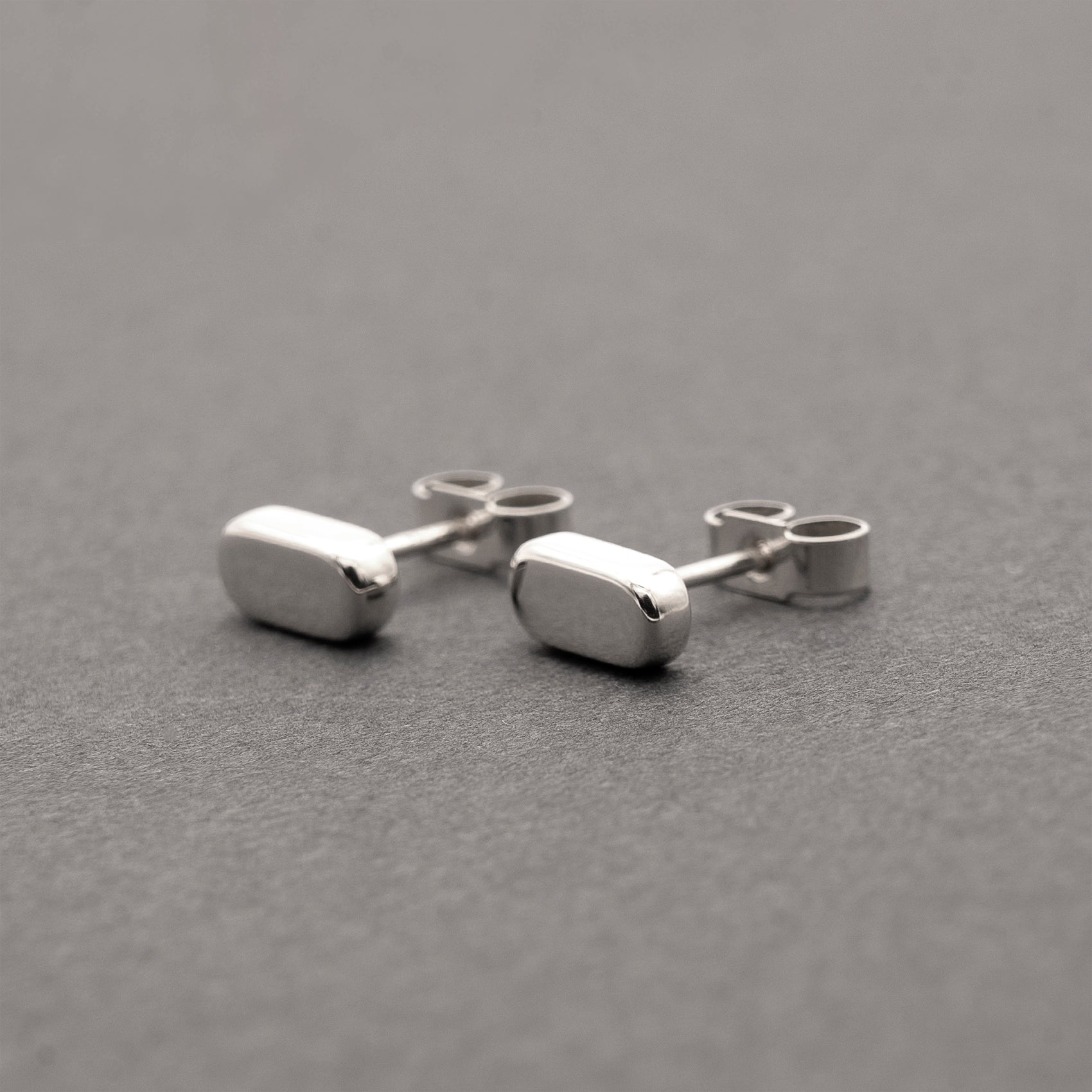 A front view of two silver earrings with a minimalist design in a flat capsule shape.