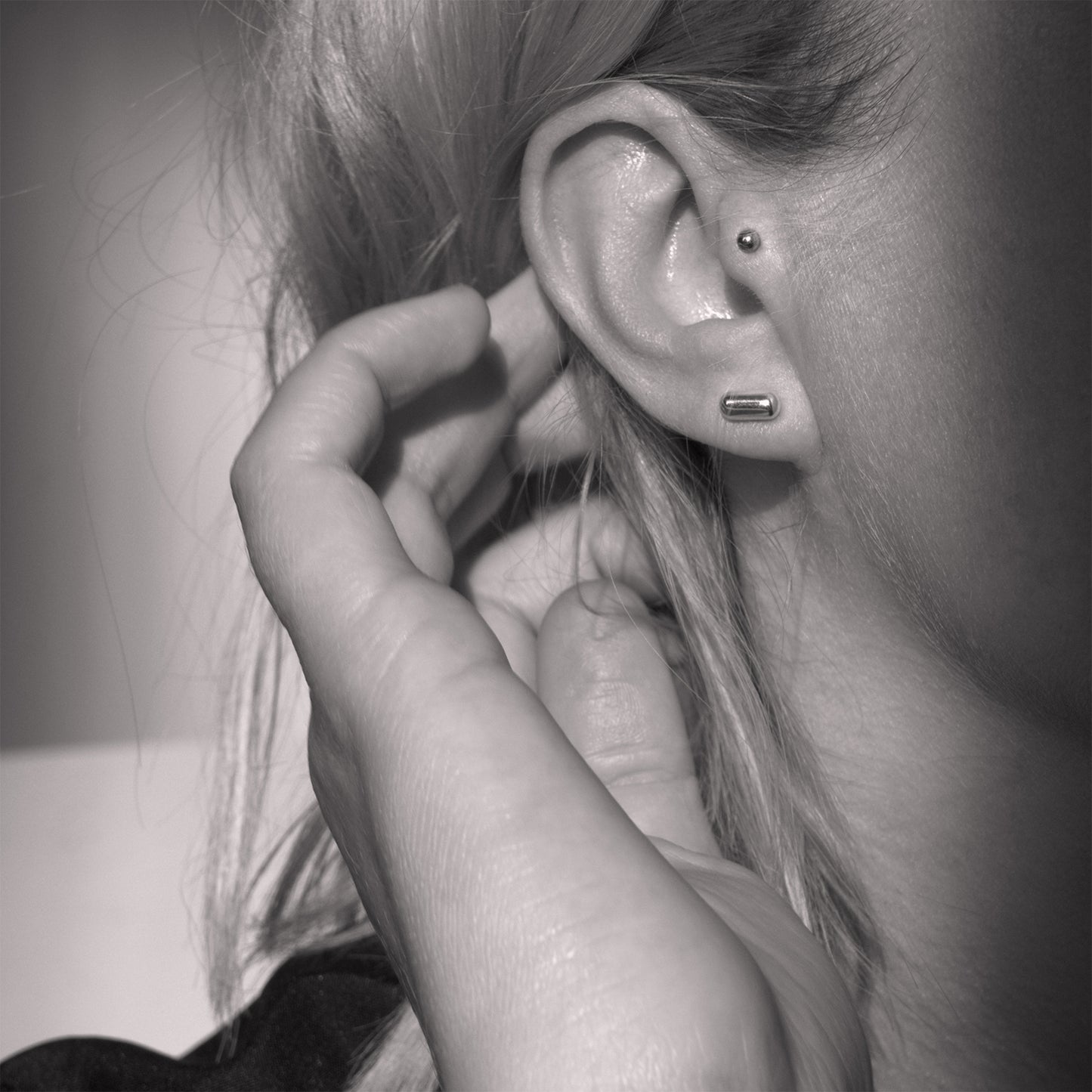 A close-up of a silver earring with a minimalist design in a capsule shape in a woman's ear. Black and white.