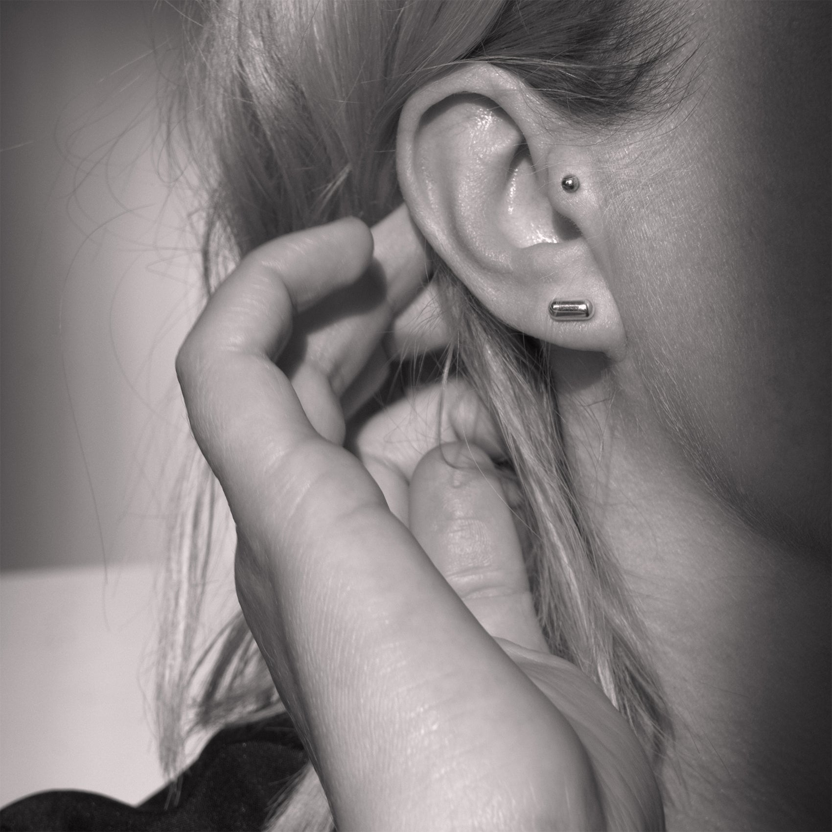 A close-up of a silver earring with a minimalist design in a capsule shape in a woman's ear. Black and white.